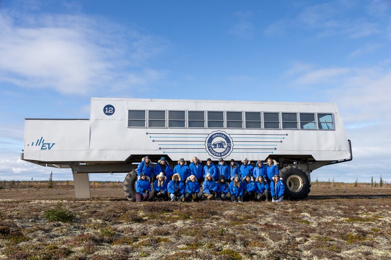 Large white EV bus with team of people in blue uniforms standing in front on tundra landscape