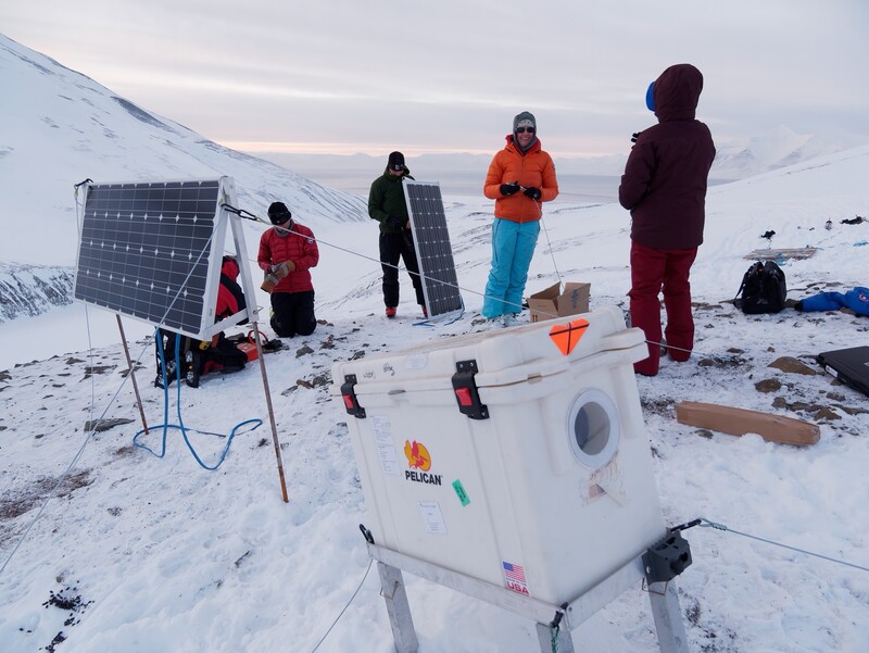 Research team with solar panel and white instrumentation box in snowy mountain environment
