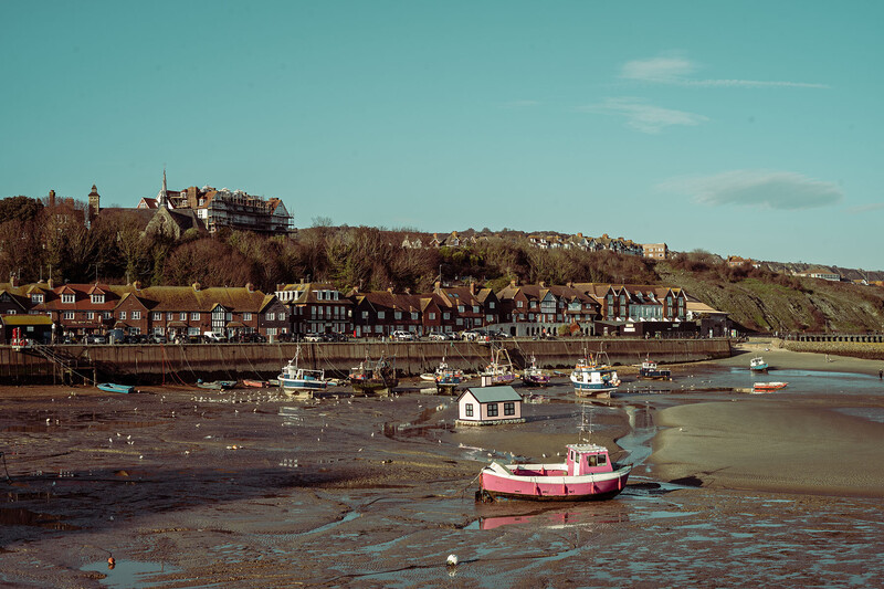 Coastal harbor at low tide with moored boats and hillside village under clear sky