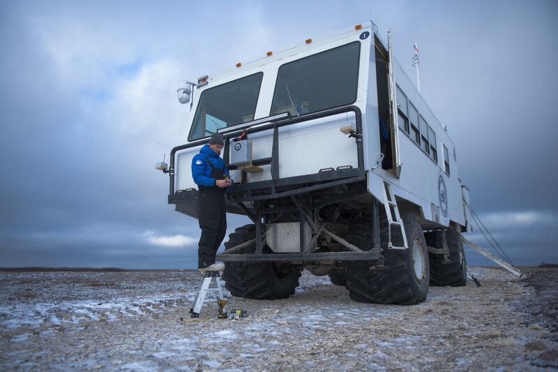 Person in blue jacket climbing ladder on side of large white expedition truck on rocky beach