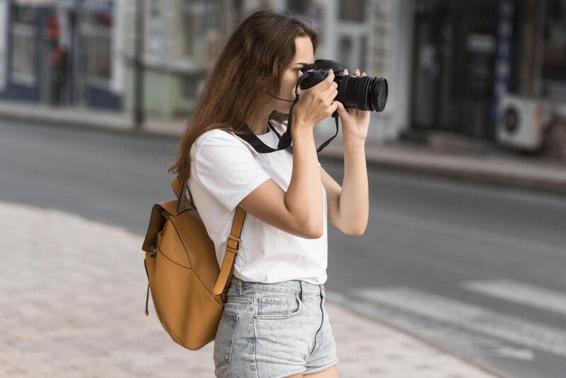 Woman with sunglasses and tan backpack photographing with DSLR camera on urban street
