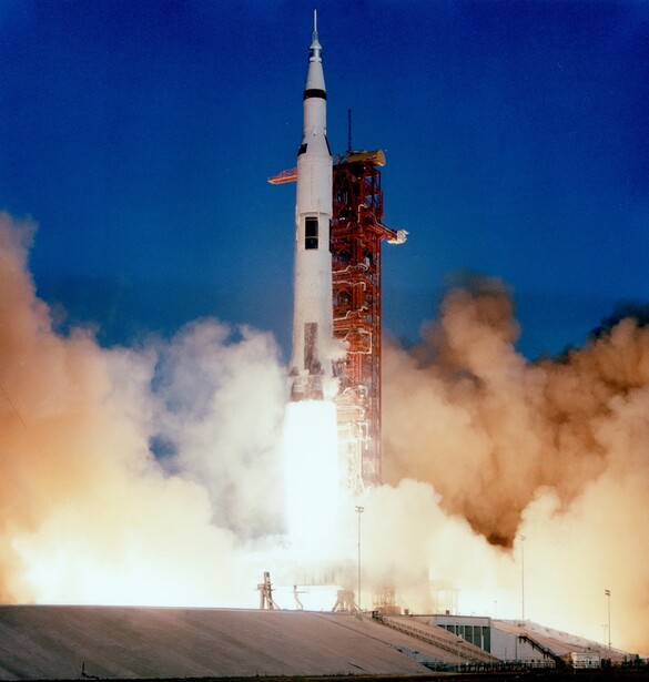 Saturn V rocket launching with massive plumes of white and tan smoke against a deep blue sky