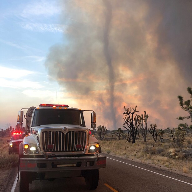 Fire truck driving toward massive wildfire with towering smoke column in desert landscape