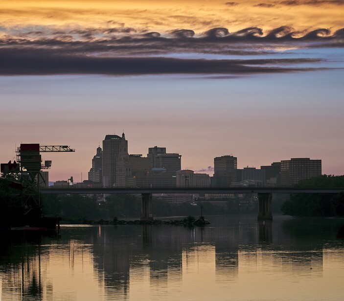 Cityscape reflected in calm water at dawn with bridge and industrial crane silhouetted against golden sky