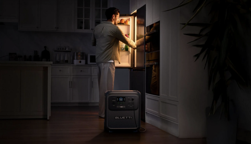 Man opening illuminated refrigerator in dark kitchen with portable power station visible