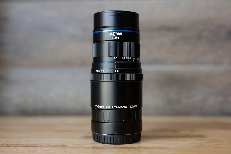 Laowa 100mm macro lens mounted vertically on wooden surface against blurred background