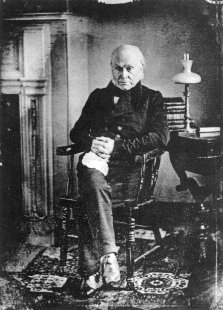 Elderly man in dark suit seated in chair holding papers, black and white portrait photograph