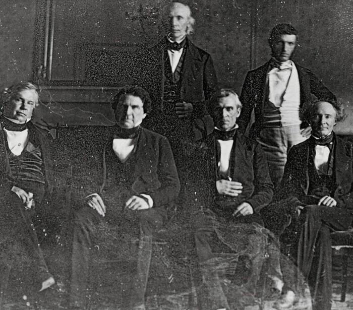Seven men in formal Victorian-era attire arranged in a group portrait