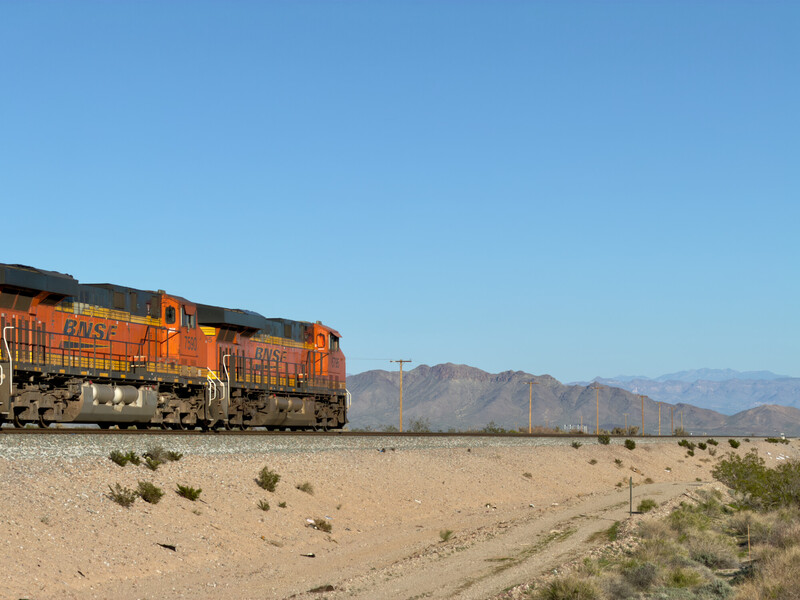 Orange and yellow diesel locomotive traveling through desert landscape with sparse vegetation and mountains in background
