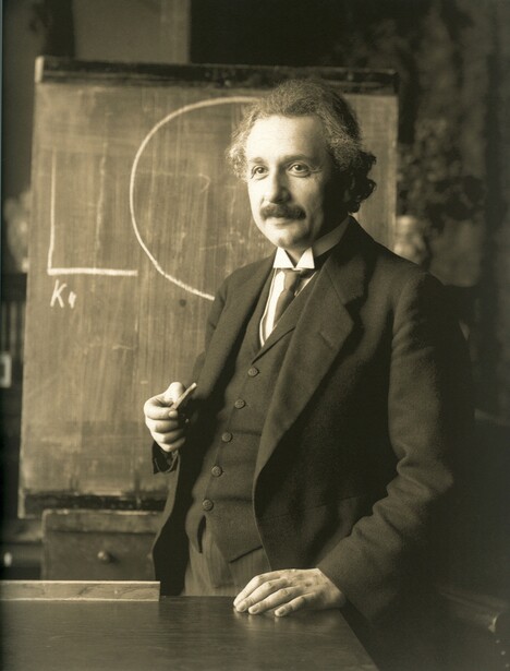 Man in dark suit and tie standing beside a blackboard with geometric diagrams