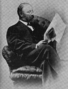 Black and white portrait photograph of a bearded man in formal attire reading a document