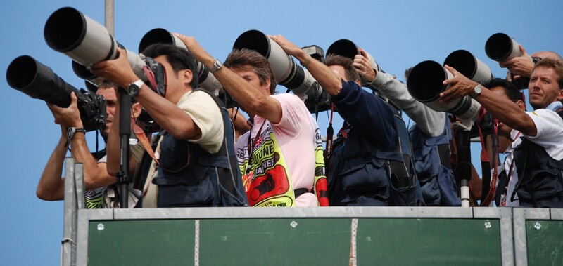 Group of photographers with telephoto lenses mounted on tripods, positioned on elevated platform against clear blue sky