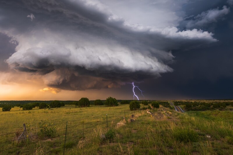 Supercell thunderstorm with anvil cloud formation and lightning bolt over rural grassland