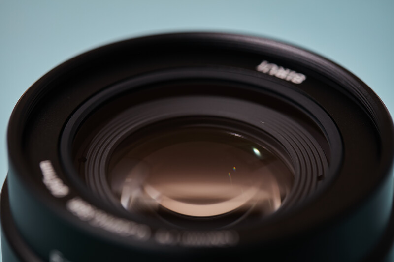 Close-up view of a camera lens front element with concentric glass rings and reflections against a light blue background