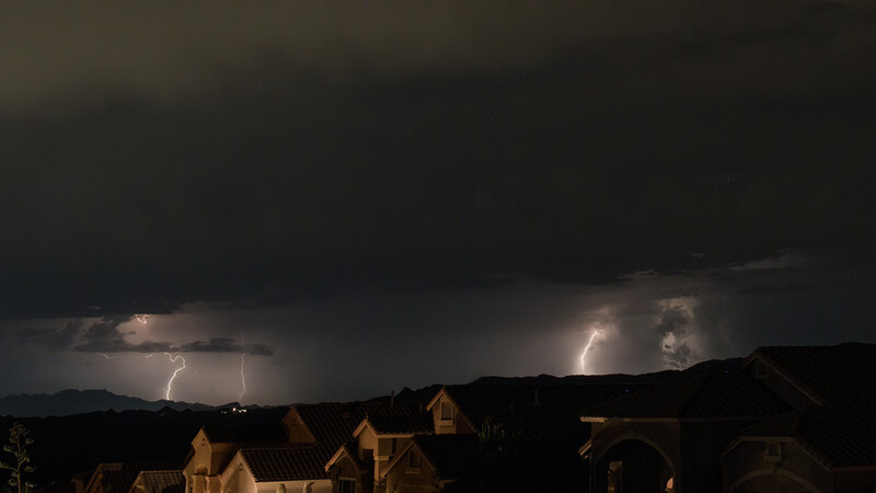 Lightning strikes illuminate dark storm clouds above residential rooftops at night