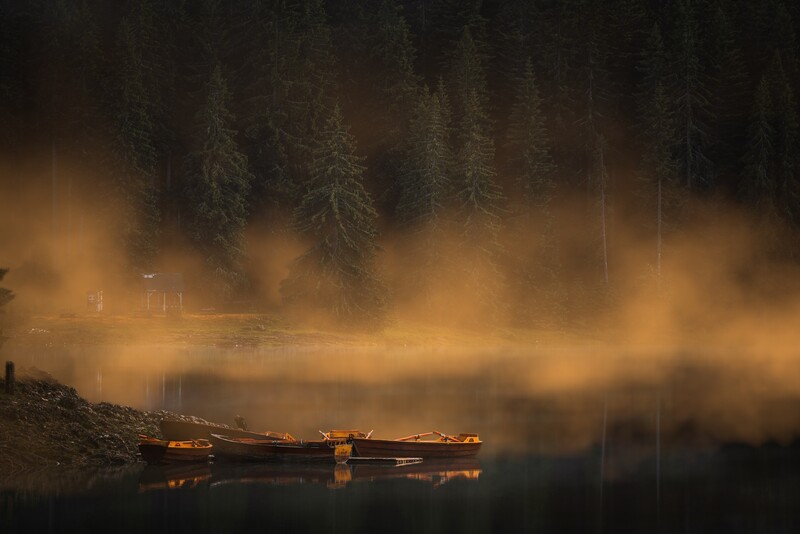 Moody forest landscape with mist rising from still water and golden boat reflections