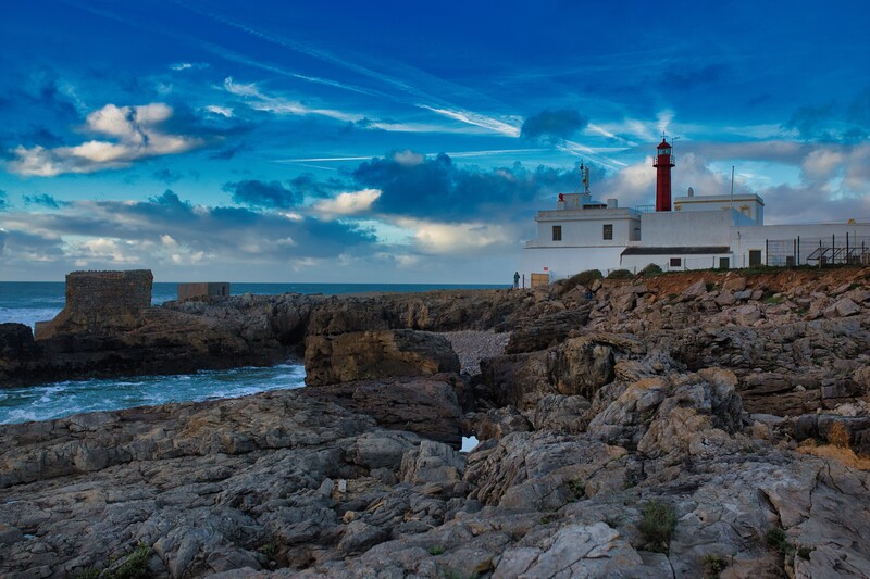 Coastal lighthouse on rocky headland under dramatic blue sky with wispy clouds