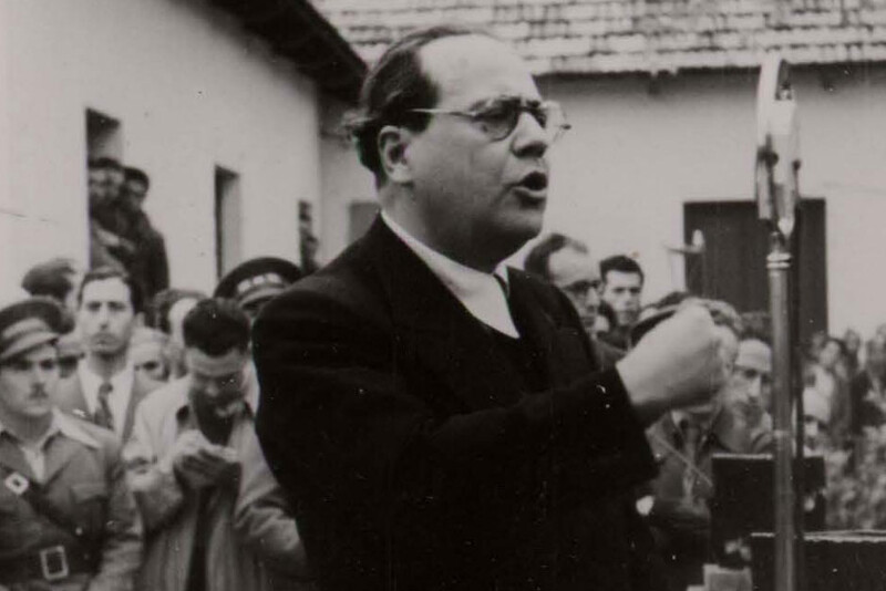 Black and white photograph of a man speaking at a microphone to a gathered crowd outdoors
