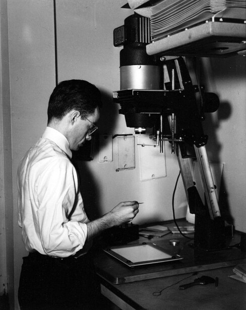 A printer works over paper on the easel beneath an enlarger, around 1950, with a wire dodging tool resting on the bench at right