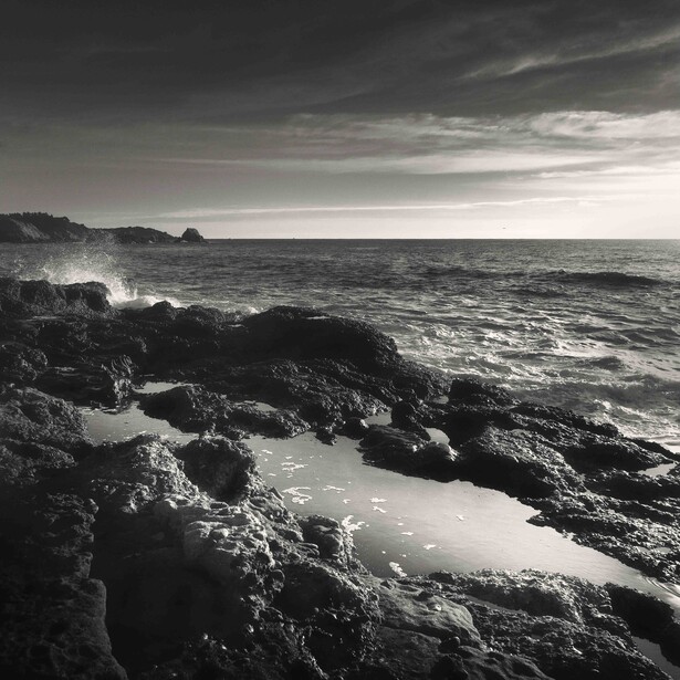 Monochrome coastal landscape with rocky foreground and calm pool reflecting light
