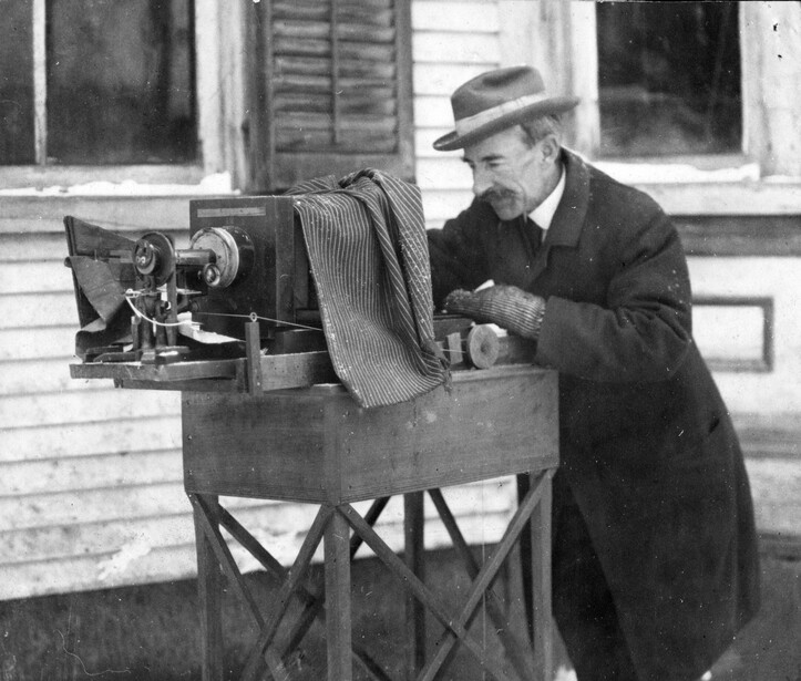Wilson Bentley bundled against the cold at his snowflake apparatus, a bellows camera mounted horizontally against a microscope on a wooden stand, the black focusing cloth draped over the back