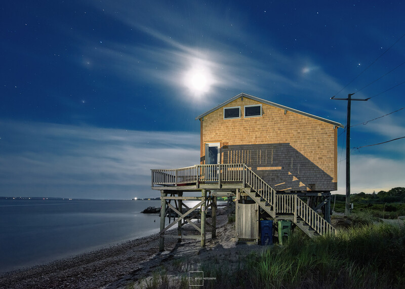 Moonlit coastal house on stilts with wooden walkway and railing at dusk