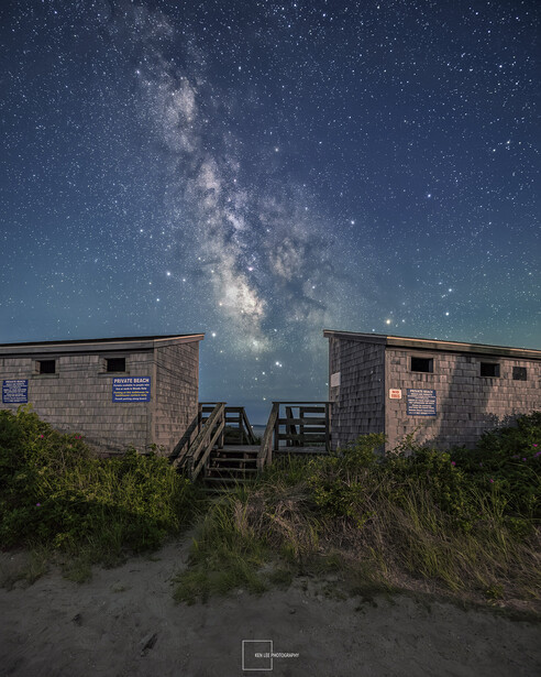 Milky Way galaxy stretches across night sky above two weathered beach houses on sand dunes