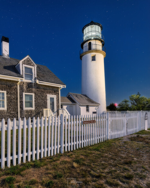 White lighthouse with black-banded top beside a shingled keeper's house with white picket fence under clear blue sky