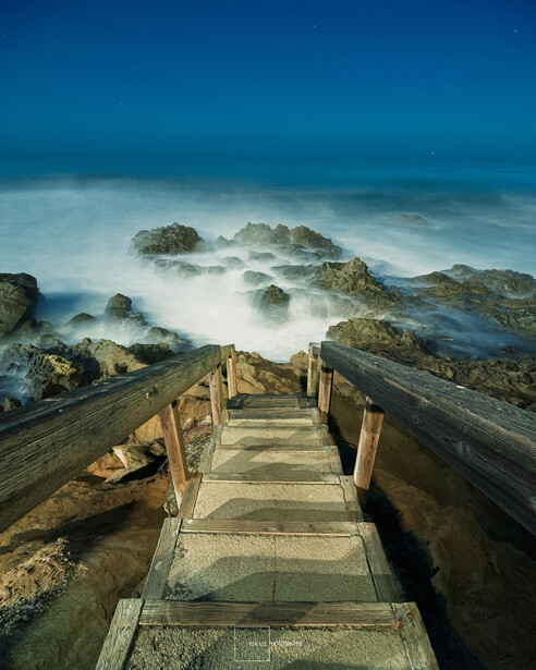 Wooden boardwalk descending toward rocky coastline with misty ocean waves and blue twilight sky