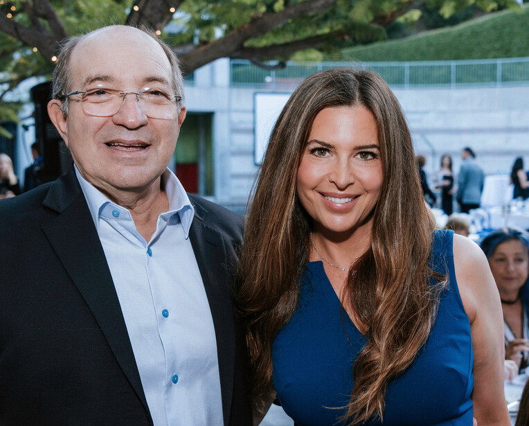 Two people posing together at an outdoor event with trees and other attendees in soft natural light