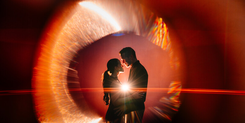 Two silhouetted figures facing each other against a glowing circular light tunnel with warm orange and white tones