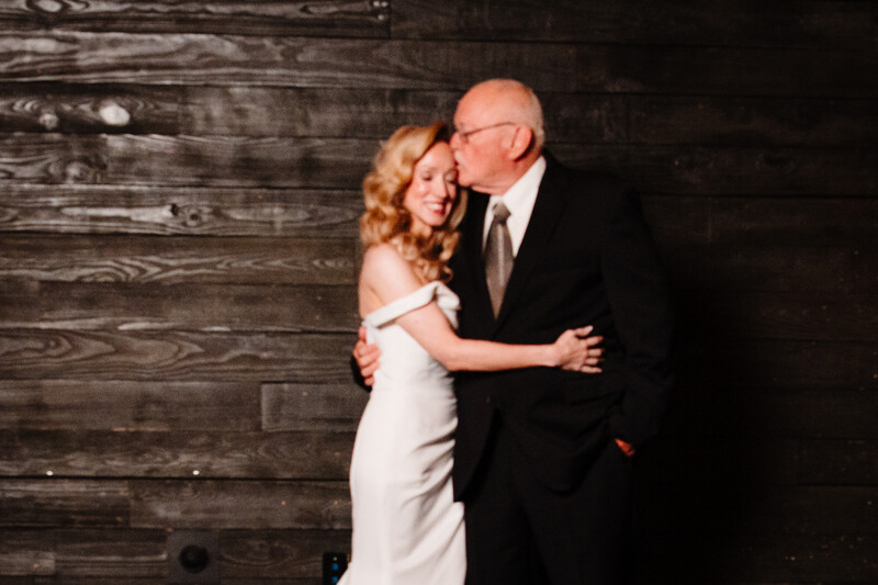 Man in dark suit kissing woman in white strapless dress against weathered wood backdrop
