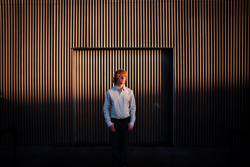 Man in light blue shirt standing centered against corrugated metal wall with dramatic side lighting