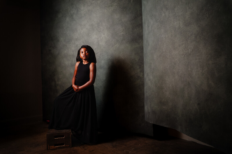 Young woman in black dress standing against textured gray walls with dramatic side lighting