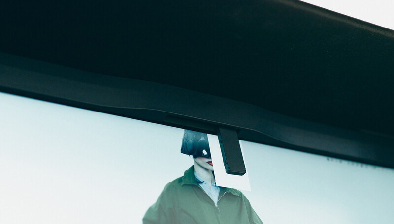 Person wearing green jacket holding large monitor above their head against white wall
