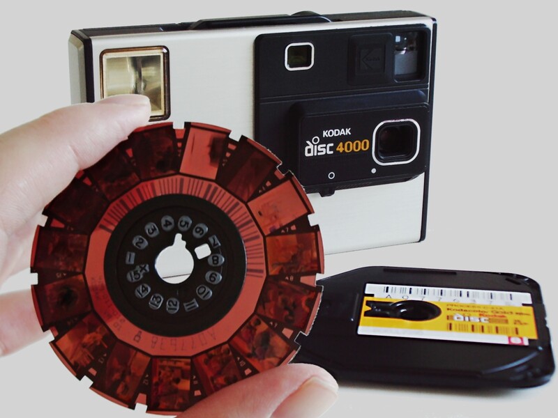 A developed disc of film held beside a Kodak Disc 4000 and its cartridge, its 15 frames arranged like numbers on a clock face around the central hub