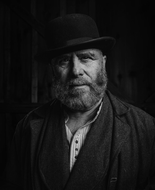 Bearded man in bowler hat and dark jacket against black background
