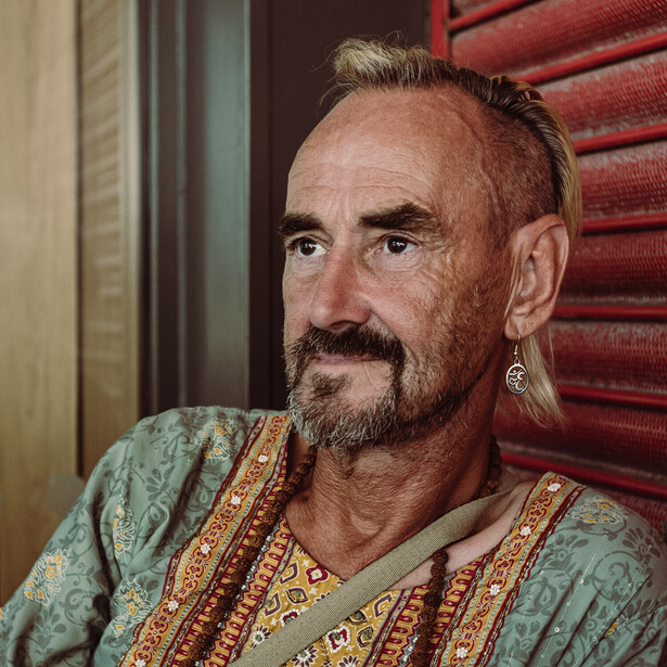 Portrait of a bearded man wearing a patterned teal and gold tunic, positioned against a red wooden background.