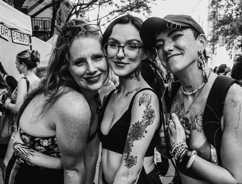 Three women with visible tattoos smiling at camera in black and white outdoor portrait