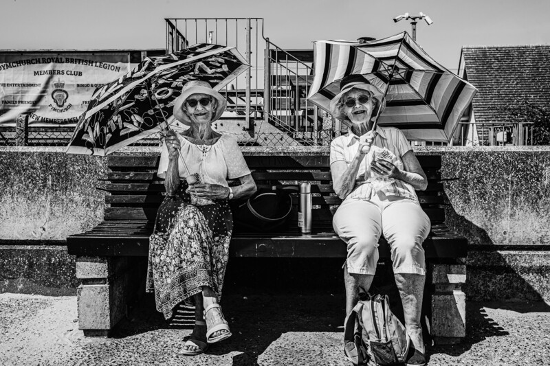 Two elderly women sitting on a bench holding umbrellas in a black and white photograph