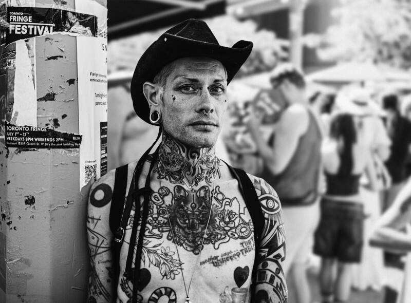 Heavily tattooed man in cowboy hat standing against festival poster in black and white