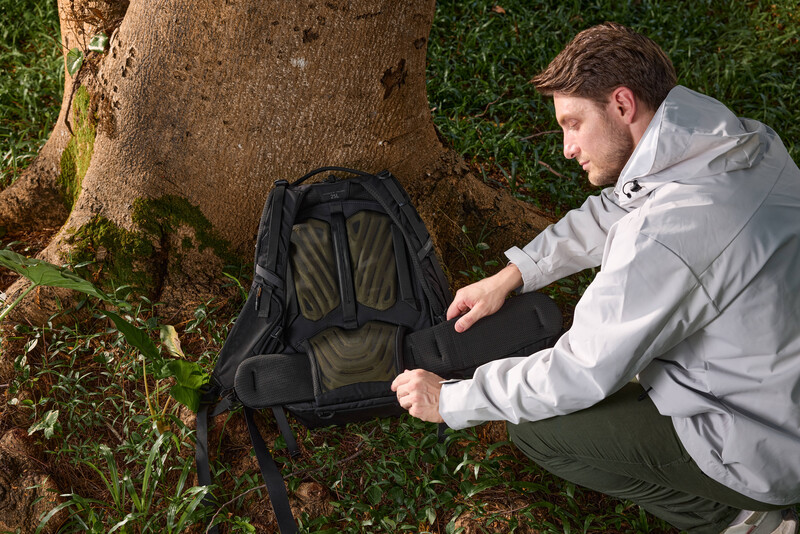 Man in light gray jacket examining a black backpack beside a large tree trunk