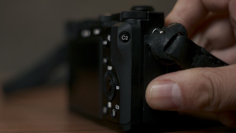 Close-up of a compact camera's rear controls being held in hands, showing C2 button and mode dial