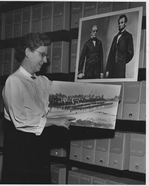 Cobb in the Still Picture stacks, holding a Brady photograph of the 20th Maine Infantry Regiment, with Brady portraits of Abraham Lincoln and Hannibal Hamlin on the shelf behind her