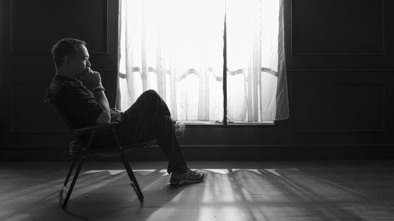 Man in dark clothing seated on chair, silhouetted against bright window with sheer curtains