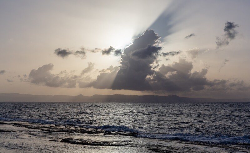 The Sun tucked behind a hard cloud edge, which is the shot the section above describes: the shafts without the disk, the highlights held rather than blown, and the beams isolated against a distant ridge and water