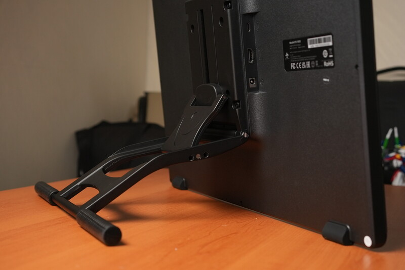 Black monitor stand with adjustable legs supporting a display on an orange desk