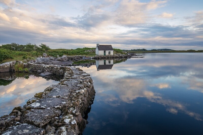 Stone boathouse reflecting in still water at golden hour with rocky shoreline