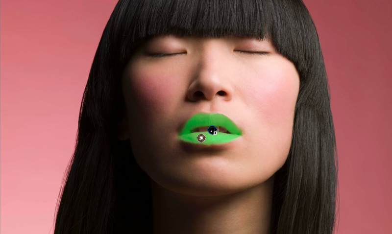 Woman with black blunt bangs and closed eyes wearing bright green lipstick against pink background