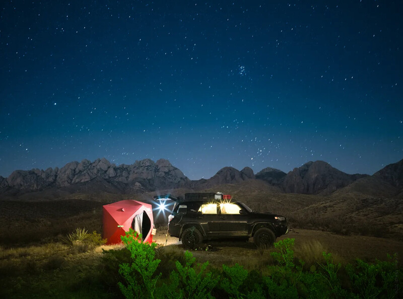 Illuminated tent and vehicle beneath starry desert sky with mountain backdrop
